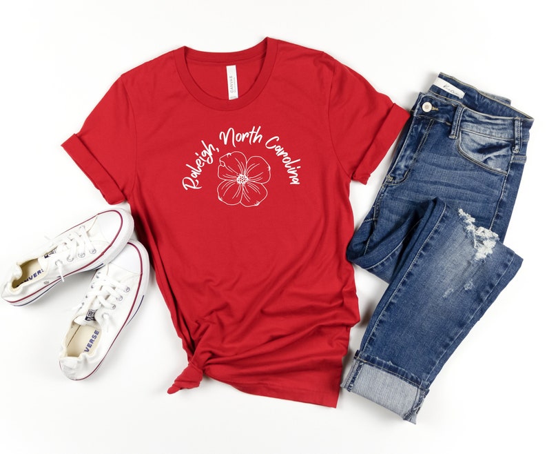 Puede incluir: Camiseta roja con texto blanco que dice "Raleigh, Carolina del Norte" y un gr&aacute;fico de flor blanca. La camiseta est&aacute; colocada sobre una superficie blanca con un par de zapatillas blancas y unos vaqueros azules.