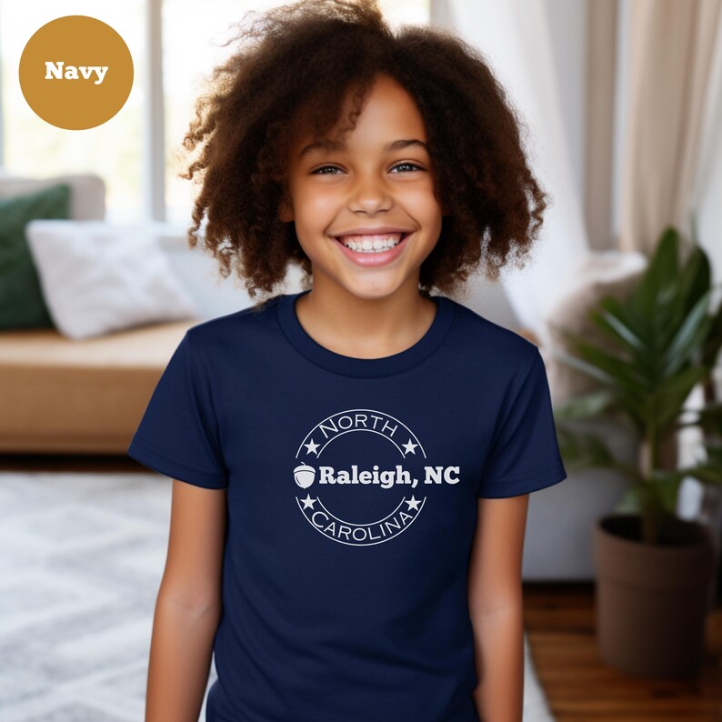 May include: A navy blue youth t-shirt with a white circle graphic featuring a stylized acorn and the text "NORTH CAROLINA Raleigh, NC".