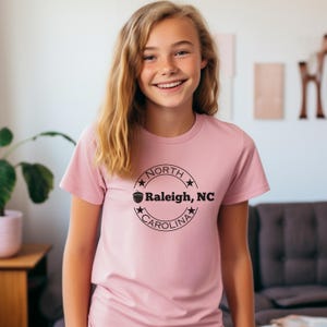 May include: Pink t-shirt with a black circle graphic featuring a stylized acorn and the text "NORTH CAROLINA Raleigh, NC".