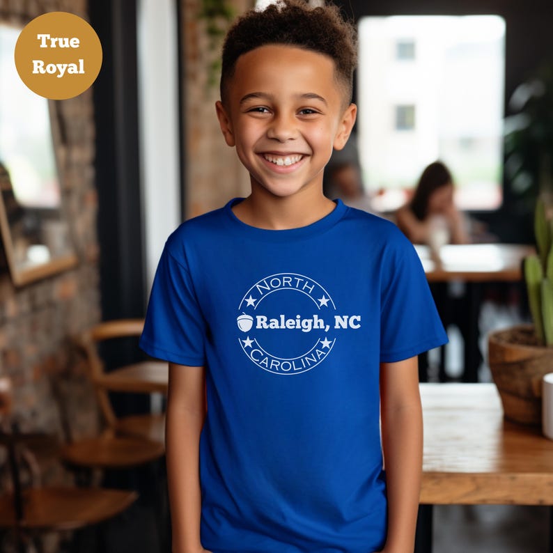 May include: A young person wearing a royal blue t-shirt with a white circle graphic on the front. The graphic features a white acorn, the words "North Carolina" and the text "Raleigh, NC".