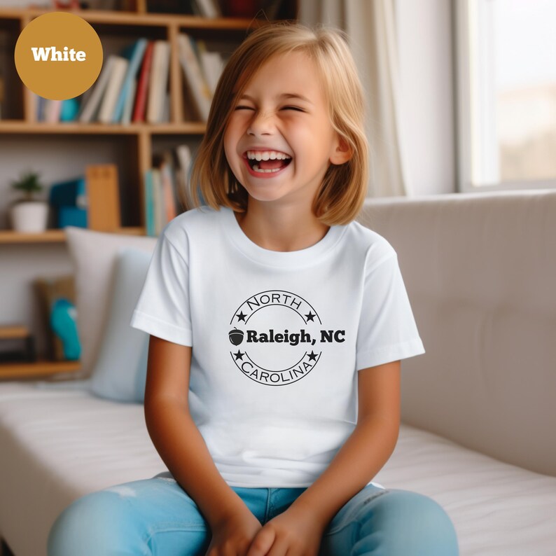 May include: White t-shirt with a black circle graphic that says "NORTH CAROLINA" with a star on each side and an acorn graphic inside the circle. The text inside the circle says "Raleigh, NC".