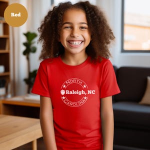 May include: A red t-shirt with a white circle design featuring the text "NORTH CAROLINA" and a stylized acorn with the text "Raleigh, NC" inside the circle.