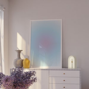 May include: A white cabinet with three drawers and gold handles sits on a wooden floor. A pink chair is in the background. A framed abstract print with a blue, pink, and white gradient hangs on the wall above the cabinet. A vase with purple flowers sits on a pink table in the foreground.
