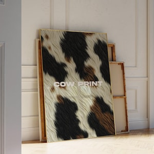 Könnte beinhalten: Ein gerahmter Leinwanddruck mit einem Kuhfellmuster in Schwarz, Braun und Creme. Der Text "COW PRINT" ist in Weiß dargestellt. Das Kunstwerk lehnt an einer Wand in einem Raum mit Holzboden.