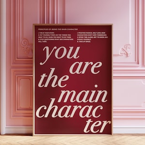 Peut inclure: Une affiche encadrée bordeaux avec les mots "you are the main character" en gras, en blanc. L'affiche comprend également une liste de principes pour être le personnage principal. L'affiche est exposée dans une pièce rose avec des panneaux muraux décoratifs.