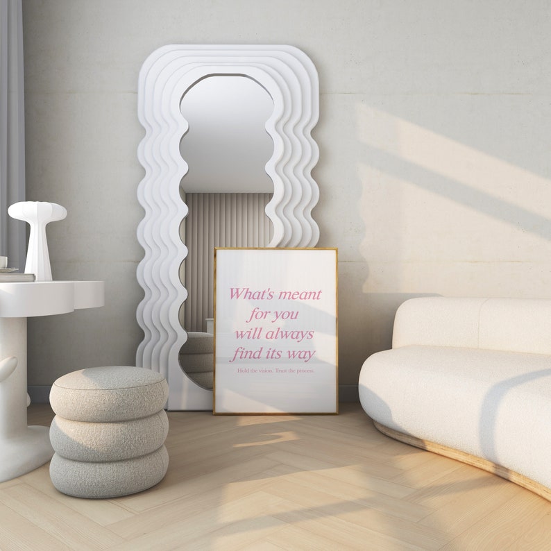 May include: A white couch with a white, wavy, framed mirror and a gold framed print with the text 'What's meant for you will always find its way. Hold the vision. Trust the process.' The print has pink text. The couch is in a room with a white floor and a white wall.
