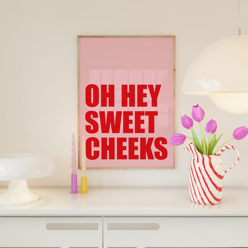 May include: A pink framed print with the text "OH HEY SWEET CHEEKS" in red letters. There are two candles and a vase with pink flowers on a white countertop.