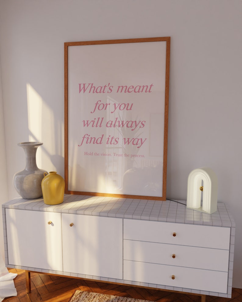 May include: A framed poster with a white background and pink text that reads 'What's meant for you will always find its way.' The poster is hanging on a white wall above a white dresser with three drawers. The dresser has gold knobs. A gray vase and a yellow vase are on the dresser. A white lamp with an arch shape is on the dresser.