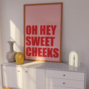 May include: A white dresser with three drawers and gold knobs. A framed poster with the text "OH HEY SWEET CHEEKS" in red letters on a pink background is on top of the dresser. A gray vase and a yellow vase are on the dresser. A white arch-shaped lamp is on the right side of the dresser.
