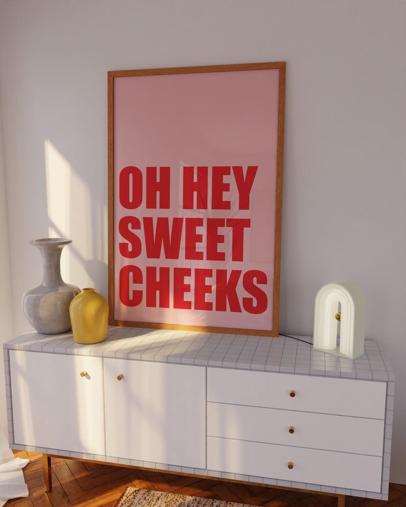 May include: A white dresser with three drawers and gold knobs. A framed poster with the text "OH HEY SWEET CHEEKS" in red letters on a pink background is on top of the dresser. A gray vase and a yellow vase are on the dresser. A white arch-shaped lamp is on the right side of the dresser.