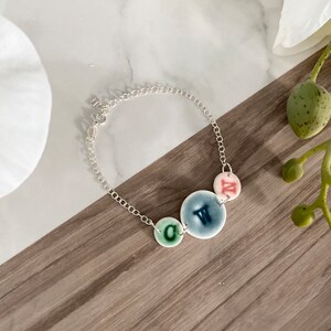 May include: A silver chain bracelet with three ceramic charms. The charms are round and have different colours and letters. The letters are C, W, and N.