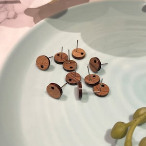 May include: A close-up of nine wooden earrings with a small hole in the centre of each. The earrings are round and have a natural wood grain pattern. The earrings are on a light blue surface.