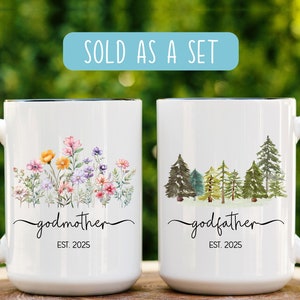 May include: Two white ceramic mugs with black handles and rims. One mug features a floral design with the word "godmother" and "EST. 2025". The other mug has a forest design with the word "godfather" and "EST. 2025". The text "SOLD AS A SET" is in a blue rectangle.