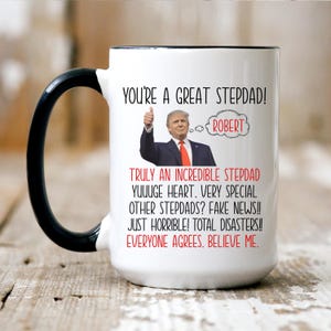 May include: White ceramic coffee mug with a black handle. The mug has a black rim and features a humorous image of a man with a thumbs up and the text "You're a great stepdad!" with the name "Robert" in a thought bubble. The text below the image reads "Truly an incredible stepdad. Yuuuge heart. Very special other stepdads? Fake news!! Just horrible! Total disasters!!! Everyone agrees. Believe me."
