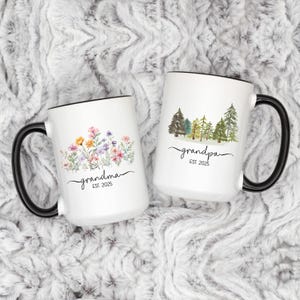 Op de afbeelding: Twee witte keramische mokken met zwarte handvatten. De mok aan de linkerkant heeft een bloemenontwerp en de tekst "grandma est. 2025". De mok aan de rechterkant heeft een bos sc&egrave;ne en de tekst "grandpa est. 2025".