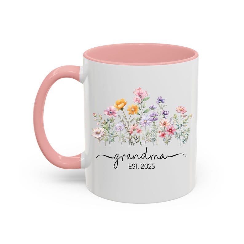 Op de afbeelding: Witte keramische mok met een roze rand en handvat. De mok heeft een aquarel-illustratie van een bloemenveld en de tekst "grandma est. 2025".