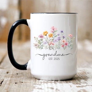 Op de afbeelding: Een witte keramische mok met een zwarte handgreep. De mok heeft een bloemenpatroon met roze, gele en paarse bloemen. De tekst "grandma EST. 2025" is op de mok gedrukt.