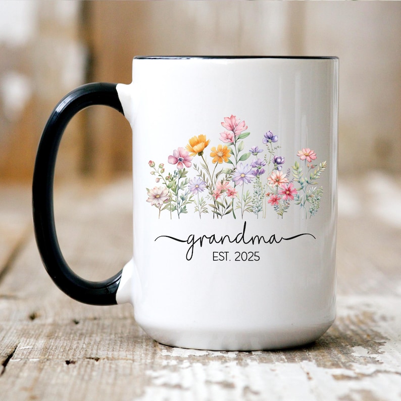 Op de afbeelding: Een witte keramische mok met een zwarte handgreep. De mok heeft een bloemenpatroon met roze, gele en paarse bloemen. De tekst "grandma EST. 2025" is op de mok gedrukt.