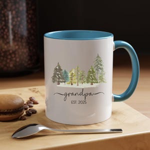 Op de afbeelding: Witte keramische mok met een blauwe rand en handvat. De mok heeft een aquarel-illustratie van een rij bomen met de tekst "grandpa EST. 2025" eronder.