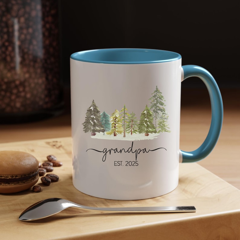 Op de afbeelding: Witte keramische mok met een blauwe rand en handvat. De mok heeft een aquarel-illustratie van een rij bomen met de tekst "grandpa EST. 2025" eronder.