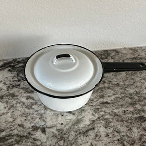 Puede incluir: Cacerola de esmalte blanco con asa y tapa negras. La tapa tiene un pomo negro.