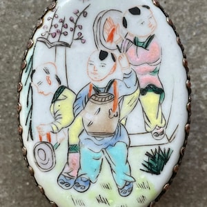 May include: An oval porcelain trinket box with a silver-toned metal rim. The box is decorated with a hand-painted scene of three children in a landscape setting. The children are wearing colorful clothing and are holding various objects. The scene is painted in a whimsical style with delicate details.