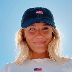Pu&ograve; includere: Un berretto da baseball in velluto a coste blu navy con una piccola bandiera americana ricamata. Il berretto &egrave; indossato con una maglietta bianca che presenta anche un piccolo emblema della bandiera americana. Lo sfondo &egrave; un cielo blu brillante.