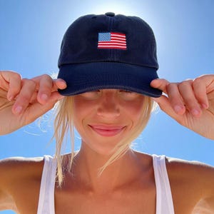 Pu&ograve; includere: Cappellino da baseball in velluto a coste blu navy con bandiera americana ricamata sul davanti. Il cappellino ha una visiera curva e un classico design a sei pannelli. L'immagine mostra anche una canotta bianca con una piccola toppa della bandiera americana.