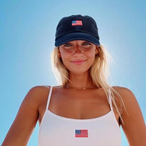 Pu&ograve; includere: Cappellino da baseball blu navy con una piccola toppa con la bandiera americana. &Egrave; visibile anche una canotta bianca a coste con una toppa con la bandiera abbinata. Lo sfondo &egrave; un cielo blu brillante.