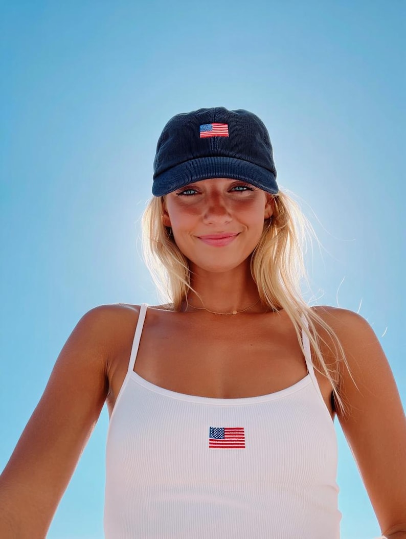 Pu&ograve; includere: Cappellino da baseball blu navy con una piccola toppa con la bandiera americana. &Egrave; visibile anche una canotta bianca a coste con una toppa con la bandiera abbinata. Lo sfondo &egrave; un cielo blu brillante.