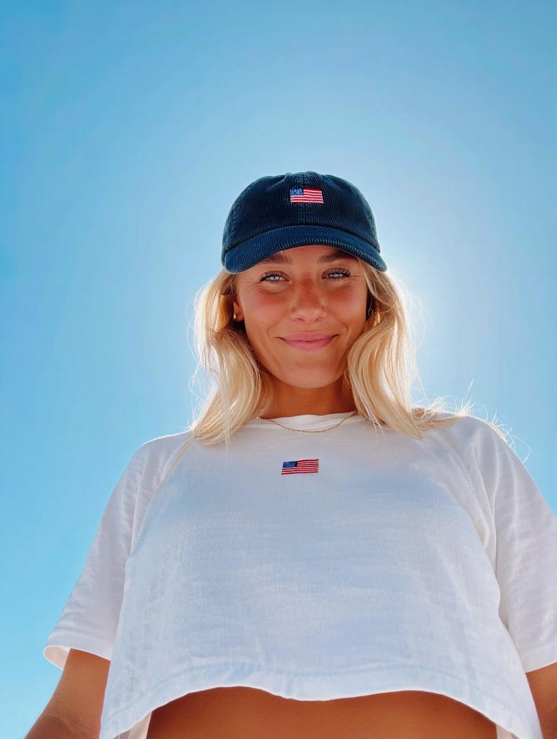 Pu&ograve; includere: Un berretto da baseball in velluto a coste blu navy con una piccola bandiera americana ricamata. Il berretto &egrave; indossato con una maglietta bianca che presenta anche un piccolo emblema della bandiera americana. Lo sfondo &egrave; un cielo blu brillante.