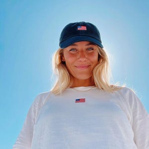 May include: A navy blue corduroy baseball cap with a small embroidered American flag patch. The cap is worn with a white t-shirt that also has a small American flag patch. The background is a bright blue sky.