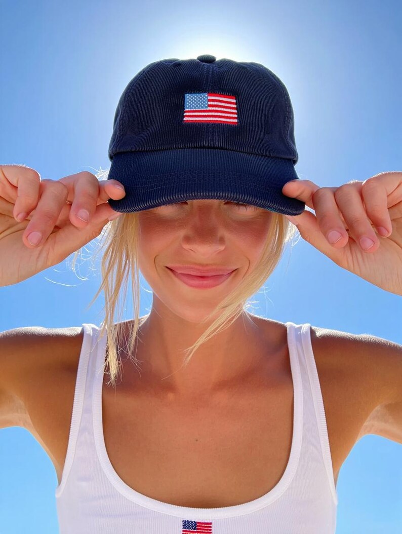 Pu&ograve; includere: Cappellino da baseball in velluto a coste blu navy con bandiera americana ricamata sul davanti. Il cappellino ha una visiera curva e un classico design a sei pannelli. L'immagine mostra anche una canotta bianca con una piccola toppa della bandiera americana.