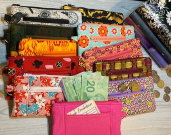 May include: A stack of colorful zippered pouches with various patterns, including floral, geometric, and abstract designs. Some pouches have a cash breakdown label and a few Canadian twenty dollar bills are visible. There are also a few coins on top of the pouches.