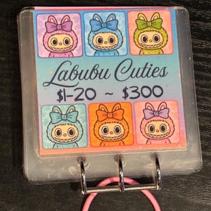 May include: A clear plastic flip-style display with a pink spiral binding. The display features six colourful squares with cartoon characters and the text "Labubu Cuties". Each character has a bow and a toothy grin.