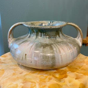 Puede incluir: Un jarrón de cerámica con dos asas, con un cuerpo redondeado y una abertura ancha. El jarrón tiene un esmalte moteado en tonos verdes, azules y blancos, con vetas rosas. La estética general es rústica y artesanal.