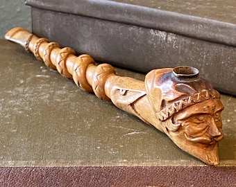 May include: A carved wooden tobacco pipe featuring a detailed Native American face and headdress. The pipe has a long, spiraled stem and a bowl with a dark, aged finish. The pipe rests on a vintage book.