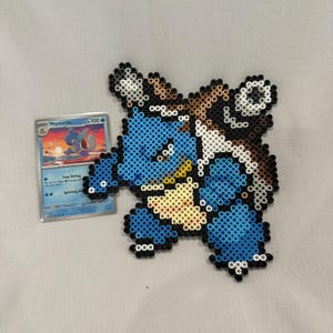 May include: A pixel art figure of the blue turtle-like creature Blastoise crafted from small plastic beads, featuring a brown shell with white cannons and a cream-colored belly. Beside it is a trading card labeled Wartortle with 100 HP.