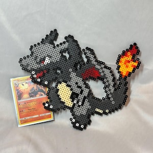May include: A gray and black pixel art dragon with red accents and a fiery tail. A Charizard Pokemon trading card is next to the dragon. The card has orange and yellow flames.
