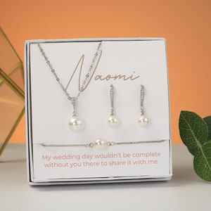 May include: A silver-coloured jewellery set comprising a necklace, earrings, and bracelet, each with a large, white pearl. The set is displayed in a white box with the name "Naomi" and the text "My wedding day wouldn't be complete without you there to share it with me."