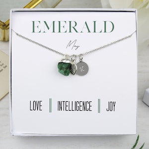 Könnte beinhalten: Eine silberne Halskette mit drei Anhängern, darunter ein roher grüner Smaragd, ein kleines Blatt und ein runder Anhänger mit einem Initial. Die Halskette wird in einer weißen Schachtel mit dem Wort "EMERALD" und dem Wort "May" präsentiert. Die Wörter "LOVE", "INTELLIGENCE" und "JOY" sind ebenfalls aufgedruckt.