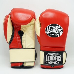 May include: Pair of red and cream-colored boxing gloves. The gloves have a cream-colored palm and wrist strap, with red on the top and sides. The wrist strap features a black and white logo that reads "PROFESSIONAL LEADERS FIGHT GEAR".