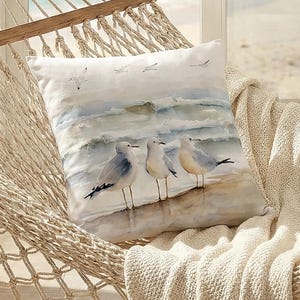 May include: Square cushion featuring a watercolour painting of seagulls on a beach. The cushion is white with a blue and grey ocean scene. It rests on a beige hammock with a cream-coloured blanket.