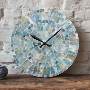 May include: Round wall clock with a mosaic design in shades of blue, white, and beige. The clock has black hands and is displayed on a wooden surface. A small blue and white decorative jar is also on the surface.