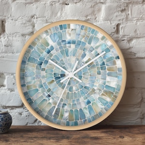 Pode incluir: Um relógio de parede redondo com um mostrador de mosaico em tons de azul, branco e bege. O relógio tem uma moldura de madeira clara e ponteiros brancos. Um pequeno vaso de cerâmica azul e branco está em uma superfície de madeira.