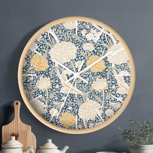 Könnte beinhalten: Eine runde Wanduhr mit hellem Holzrahmen. Das Zifferblatt zeigt ein Blumenmuster in Blau-, Creme- und Gelbtönen. Weiße Zeiger zeigen die Uhrzeit an. Die Uhr ist an einer dunkelblauen Wand befestigt.