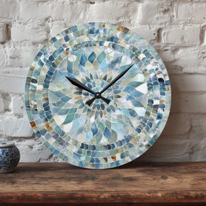 May include: Round wall clock with a mosaic design in shades of blue, teal, and white. The clock face features a floral pattern and black clock hands. The clock is displayed on a wooden surface against a white brick wall.