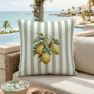Puede incluir: Un cojín decorativo con un diseño de rayas verdes y blancas. Presenta una ilustración de acuarela de limones en una rama con hojas verdes. El cojín está sobre un sofá beige al aire libre.