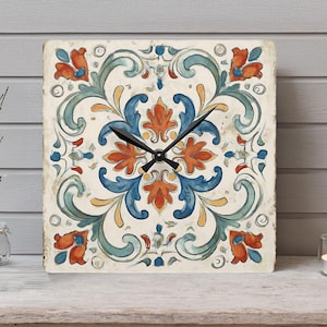 May include: Square wall clock with a vintage, floral tile design. The clock face features a cream background with orange, blue, and green floral patterns. Black clock hands indicate the time. The clock is displayed on a wooden shelf.