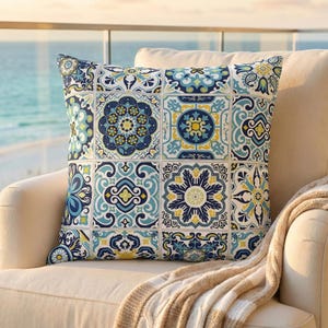 Puede incluir: Cojín cuadrado con un diseño de azulejos en tonos azules, amarillos y blancos. El cojín está colocado sobre una silla color crema con una manta beige. El fondo muestra una vista del mar y el cielo.